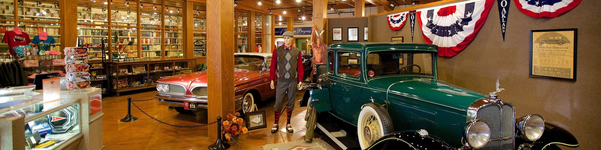 Exposição de carros antigos com um carro verde e um carro vermelho em exibição, cercada por estantes de livros e bandeiras americanas.