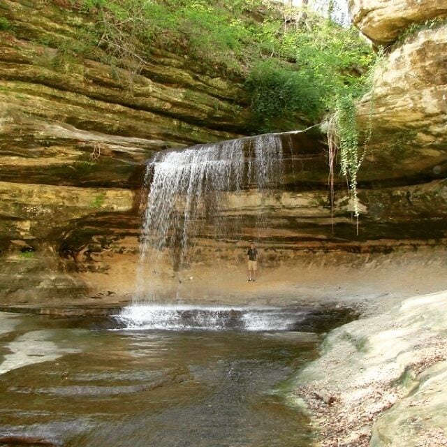 A waterfall cascading over a rocky cliff into a shallow pool surrounded by layered rock formations.