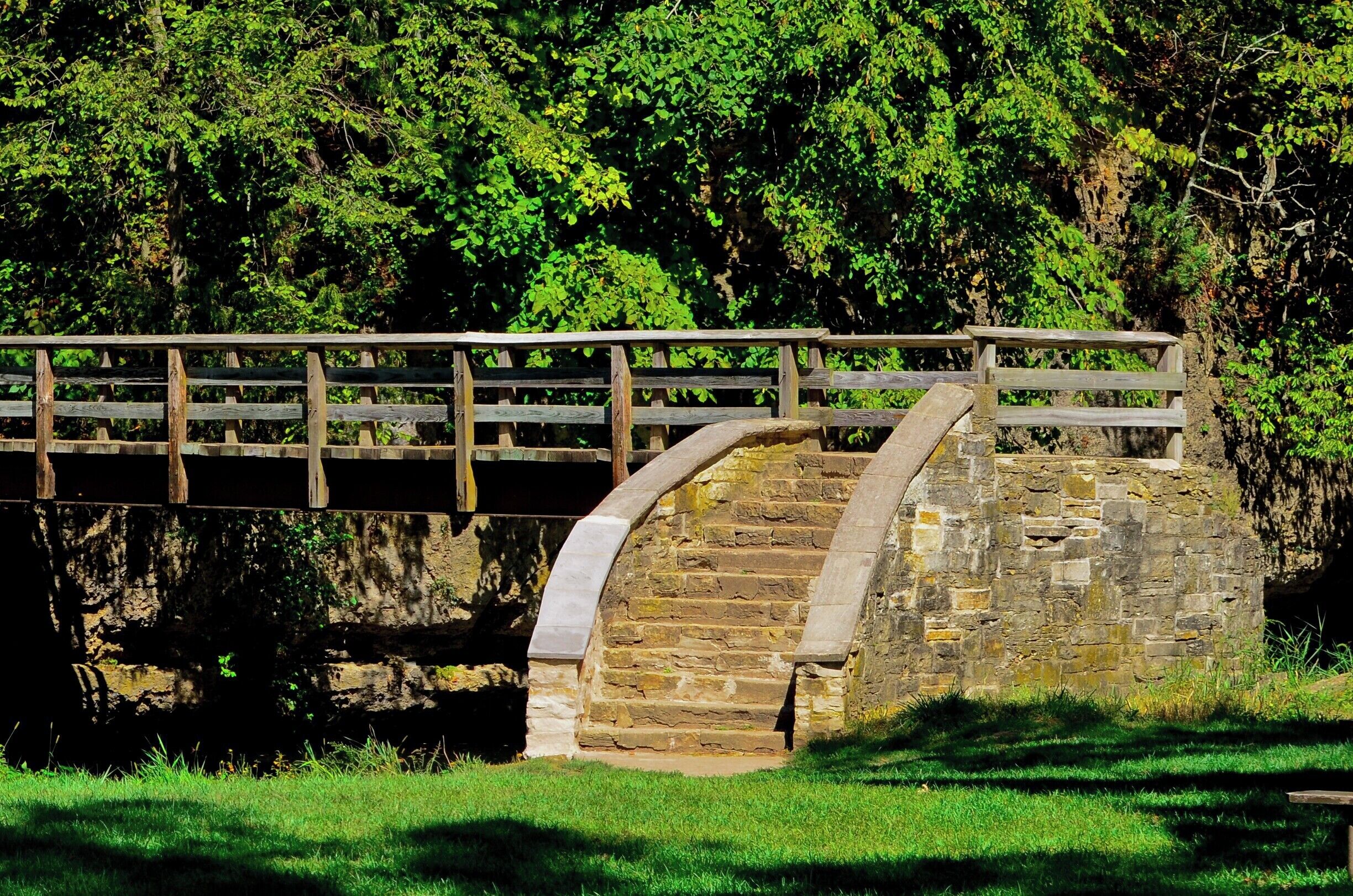 A stone bridge with a wooden railing over a river, surrounded by lush greenery.
