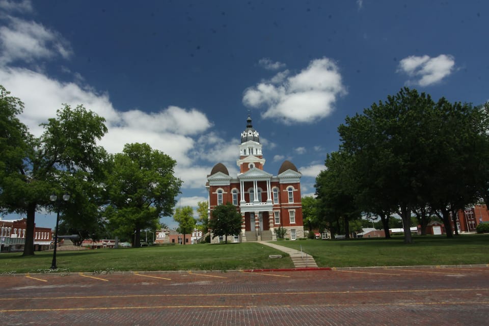 A historic red brick building with a central dome and a clock tower, surrounded by green trees and a clear blue sky.