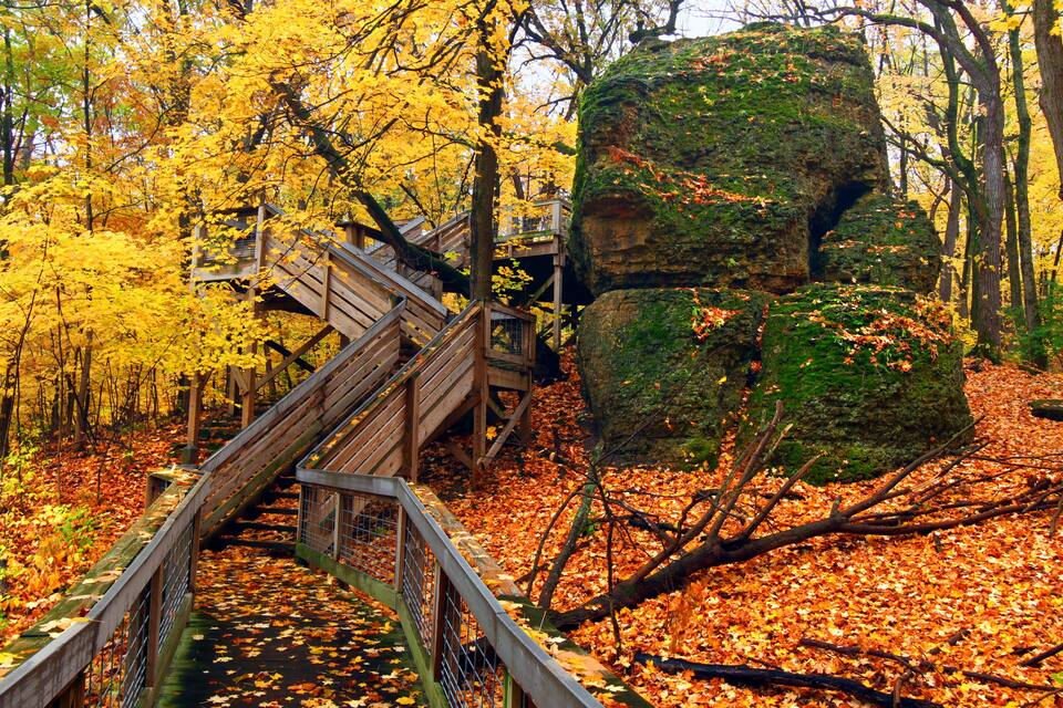 A wooden bridge over a forest with yellow leaves and a large rock formation.