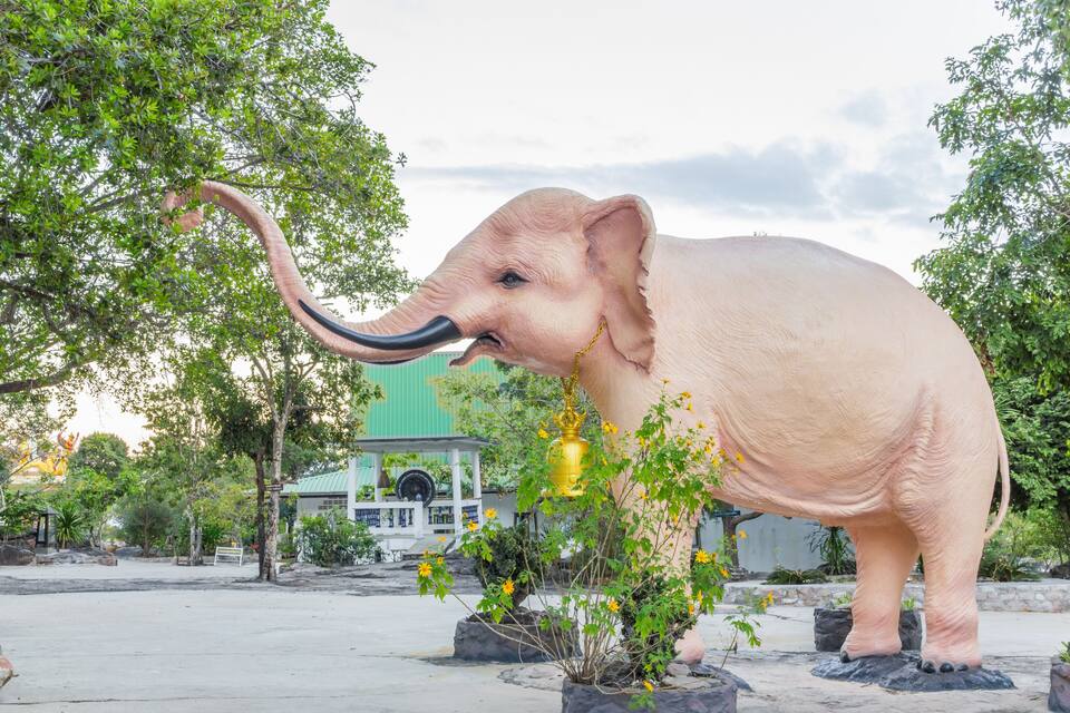A large elephant statue in a park with trees and a building in the background.