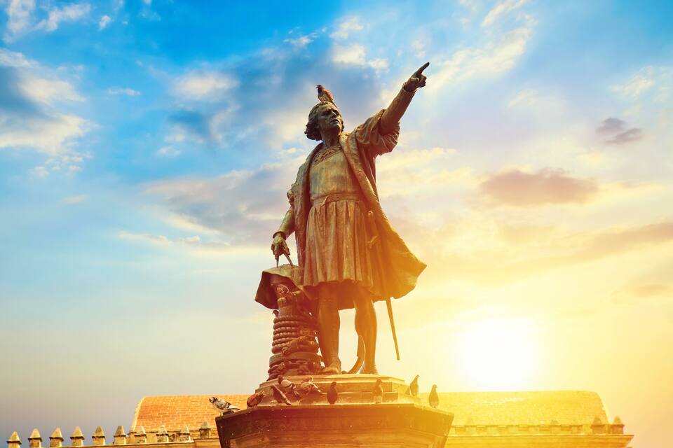 A statue of a historical figure with a bird on their shoulder, pointing upwards, with a bright sun in the background.
