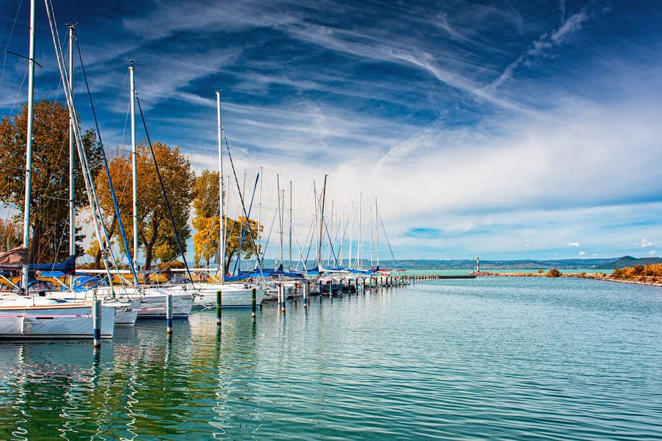 Un port de plaisance où sont amarrés de nombreux voiliers le long du rivage, entouré d’arbres et avec une côte lointaine en toile de fond.