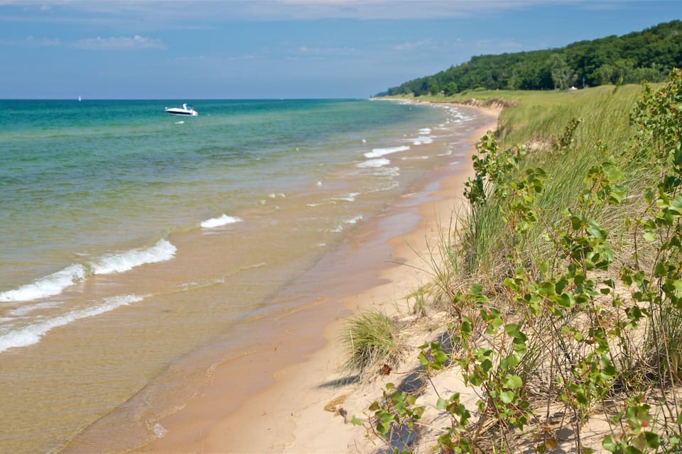 A sandy beach with gentle waves, a boat in the water, and a forested hill in the background.