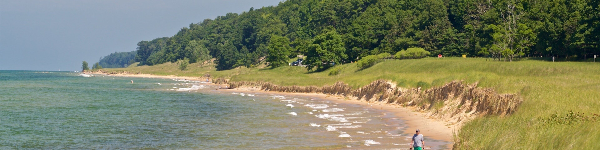 Ein Strand mit sanften Wellen, eine Person geht spazieren und ein bewaldeter Hügel im Hintergrund.