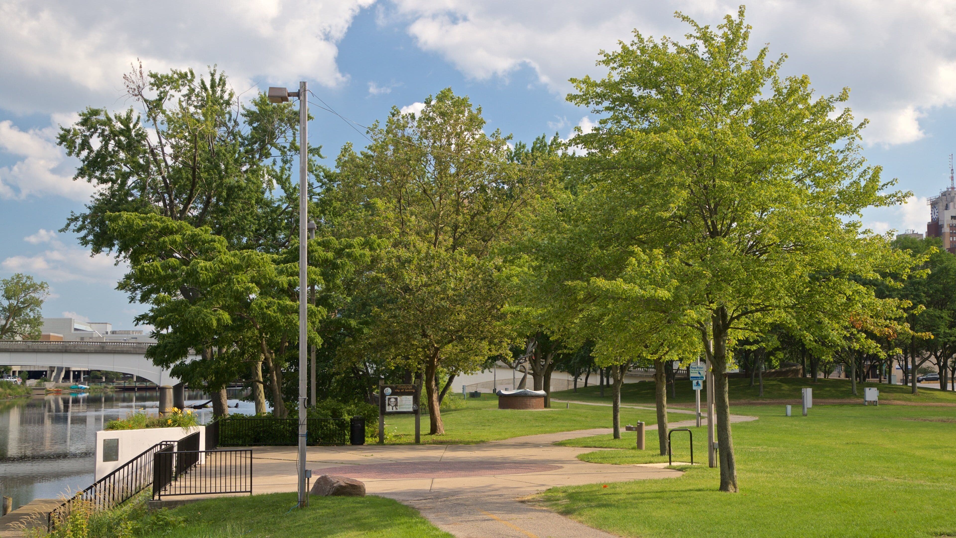 A park with a river, trees, a bridge, and a pathway.