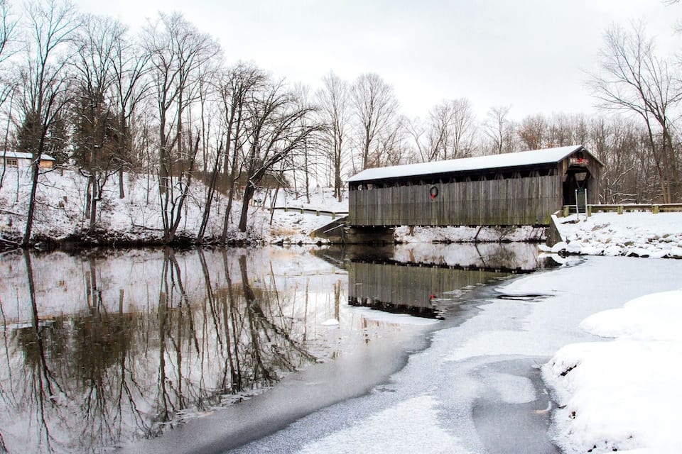 Un puente cubierto de madera sobre un río helado, rodeado de árboles cubiertos de nieve y un pequeño edificio.