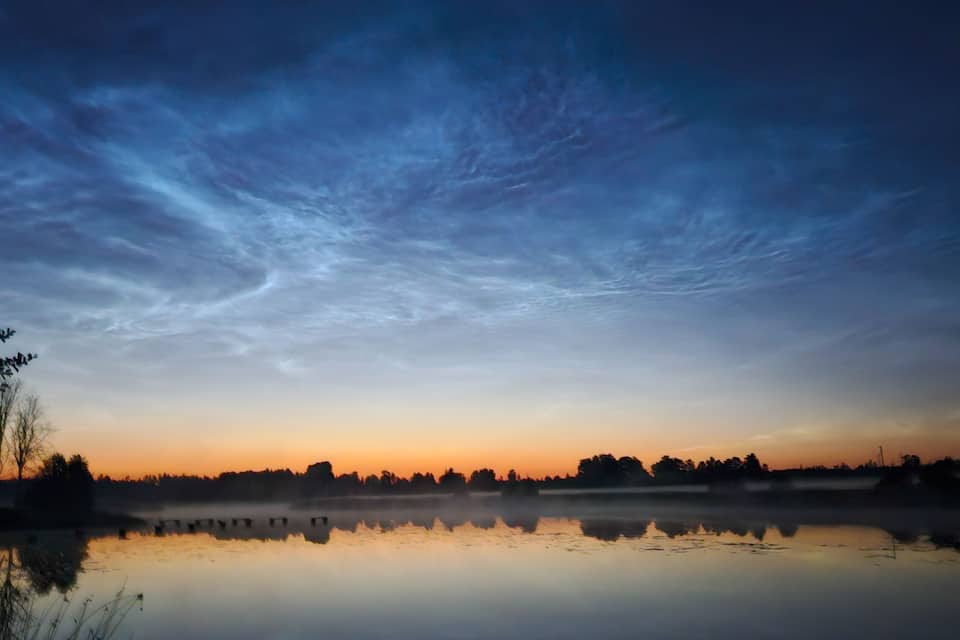 A calm lake at sunrise with a clear sky and silhouettes of trees and structures on the horizon.