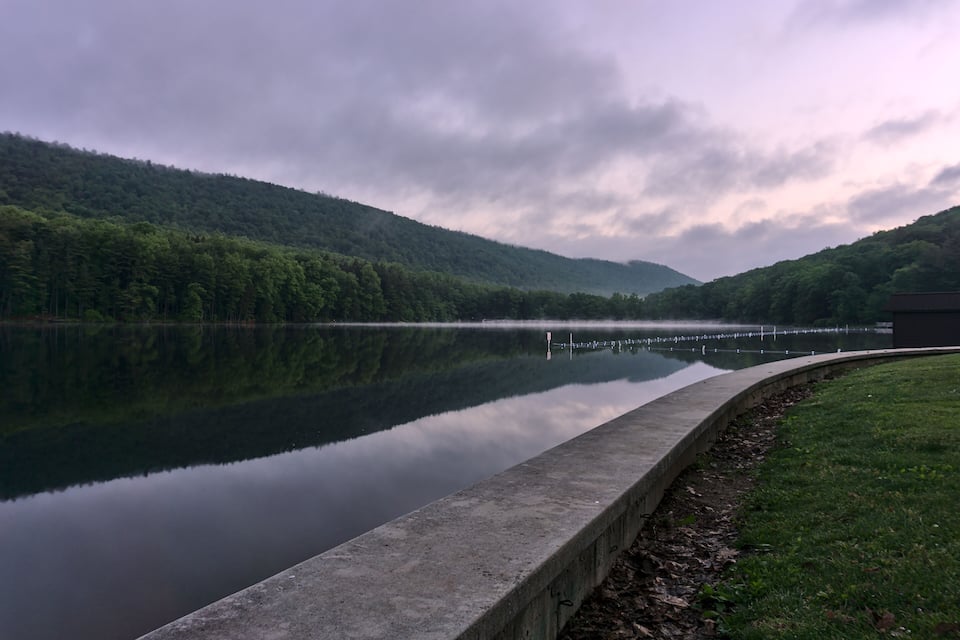 A calm lake with a concrete embankment, surrounded by lush green hills under a cloudy sky.