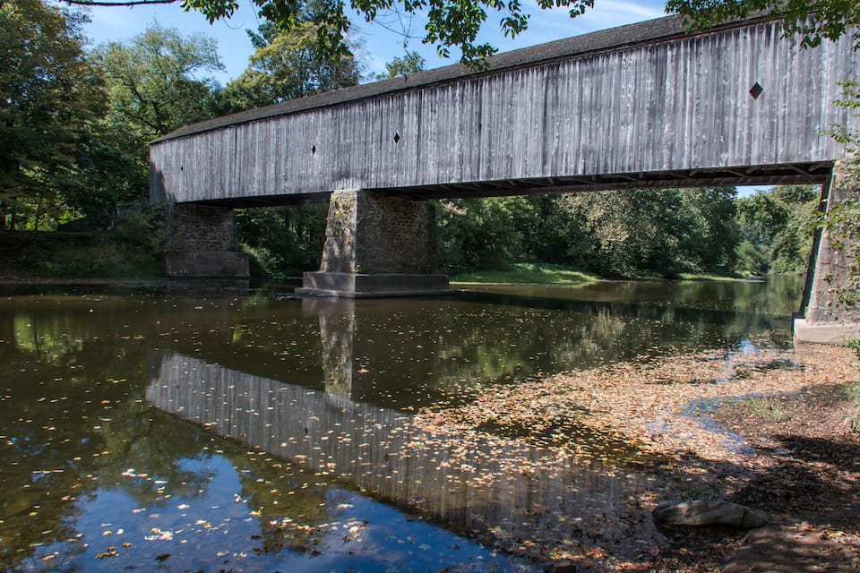 A wooden covered bridge over a calm river with reflections of trees and leaves on the water.