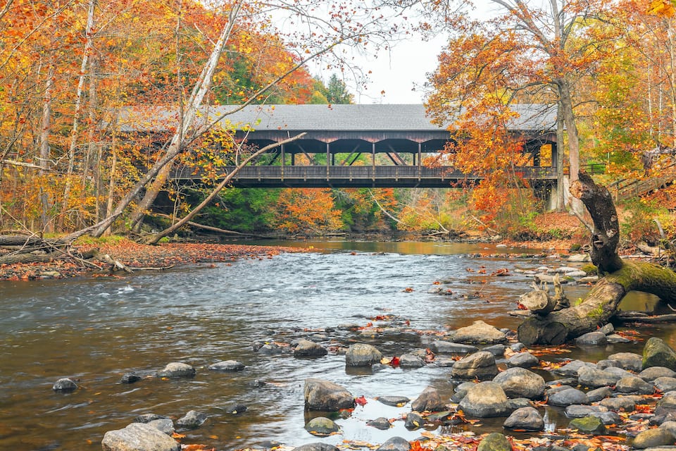 A river with rocks and autumn leaves, a wooden bridge, and a building with a covered walkway.