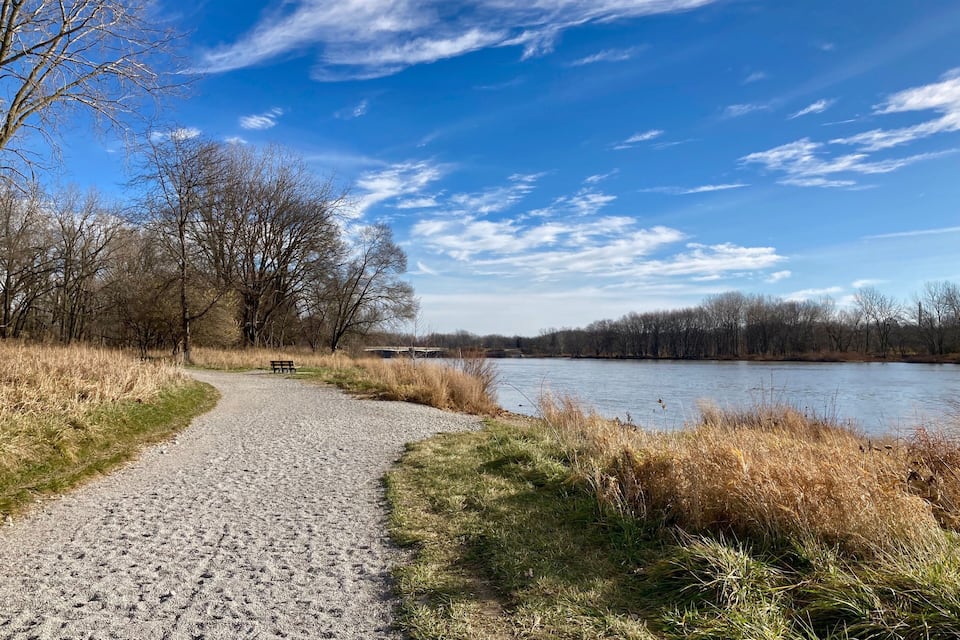 Un sendero de grava que conduce a un banco junto a un río, rodeado de árboles y zonas verdes.