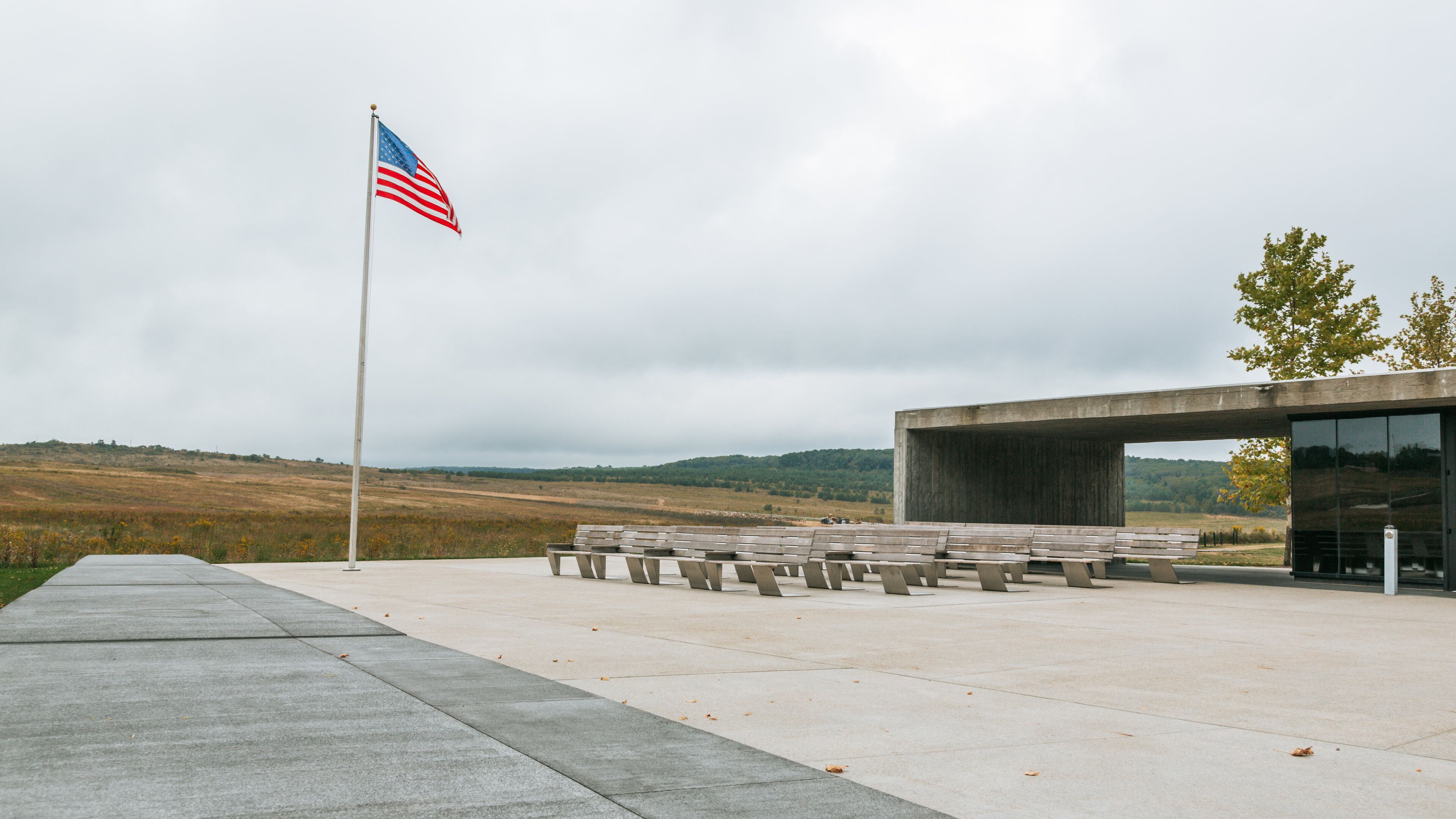 A modern building with benches, an American flag, and a hilly landscape in the background.
