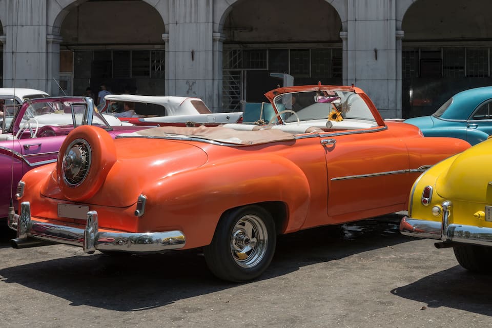 Un coche descapotable naranja antiguo estacionado en un estacionamiento junto a otros coches clásicos.