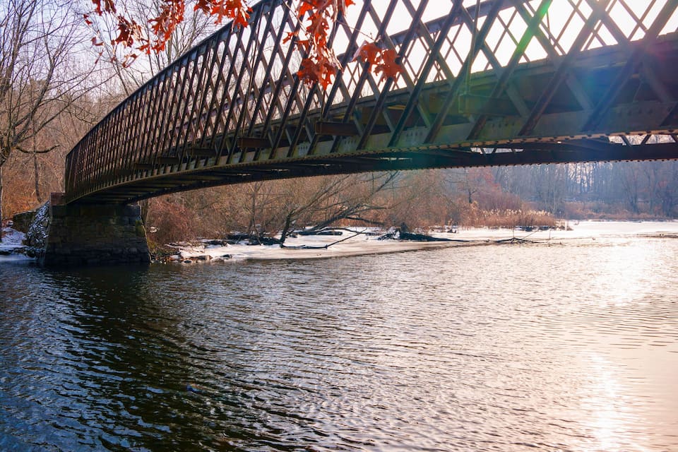 Un pont d’acier enjambant une rivière, avec des feuilles d’automne et de la neige sur le sol.