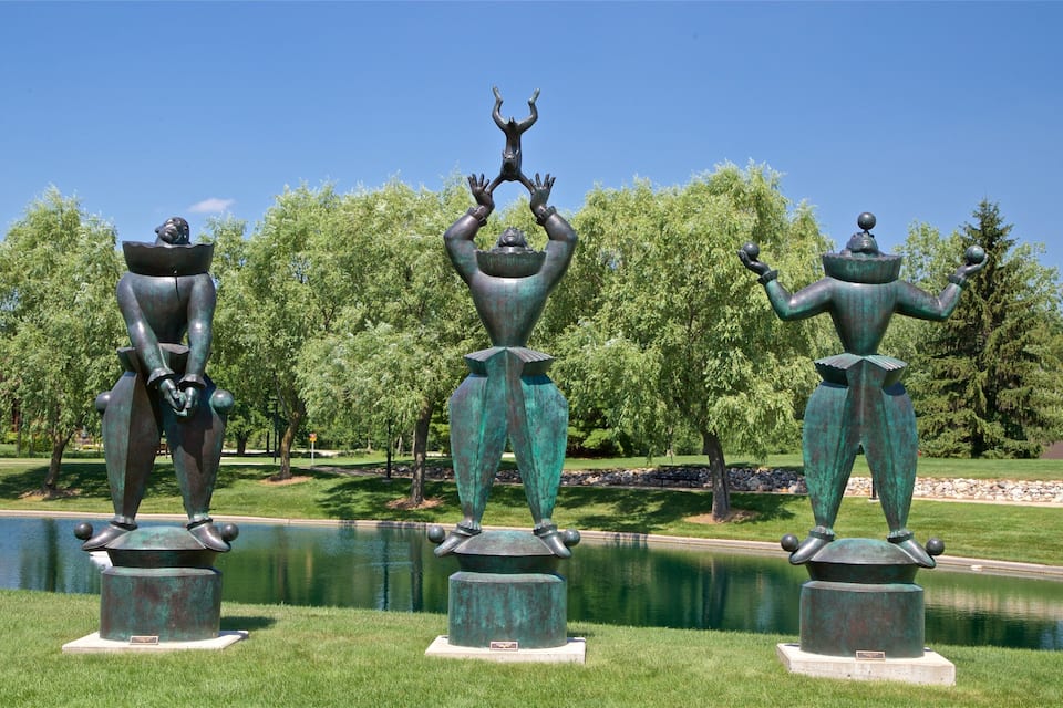 Drie bronzen beelden in een park, met een vijver en bomen op de achtergrond.