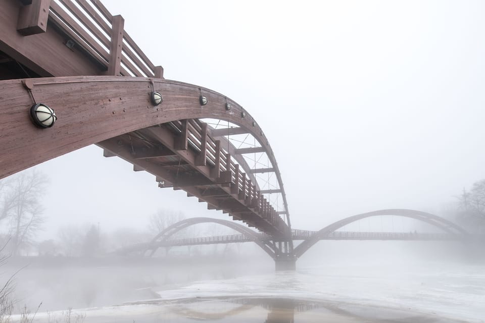 A wooden bridge with a curved arch over a river, surrounded by fog and bare trees.