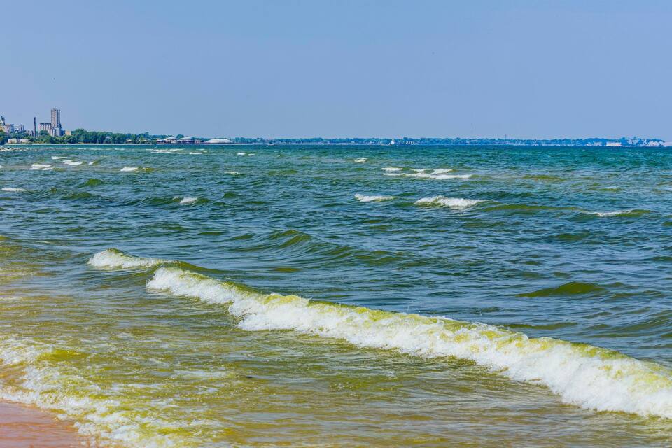 A coastal view with waves, a sandy beach, and a distant industrial area.