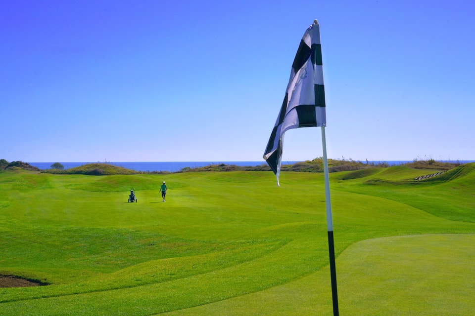 Un campo de golf con una bandera, un asta y una vista distante del océano.