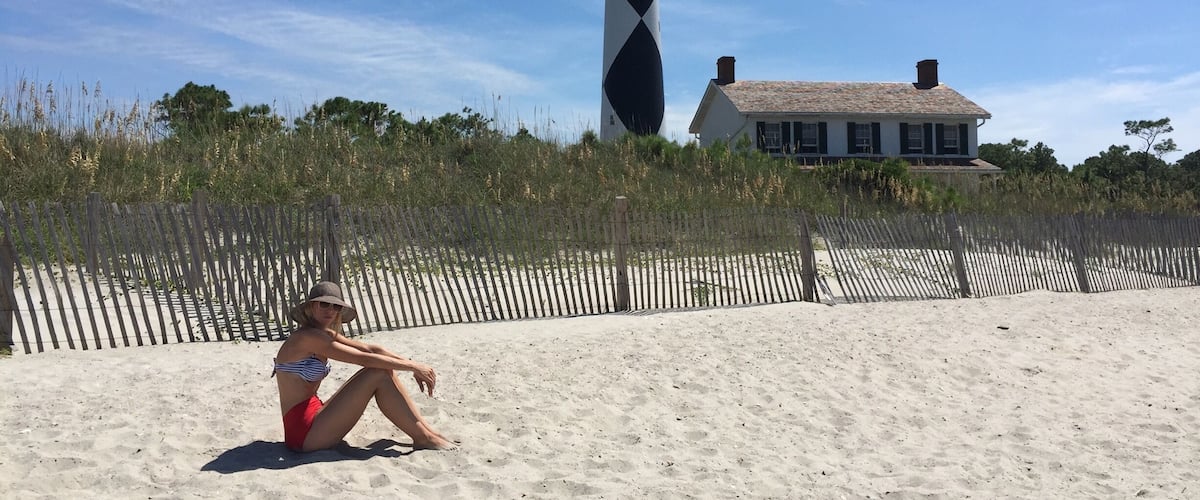 A beach with a person sitting on the sand, a lighthouse, and a house in the background.