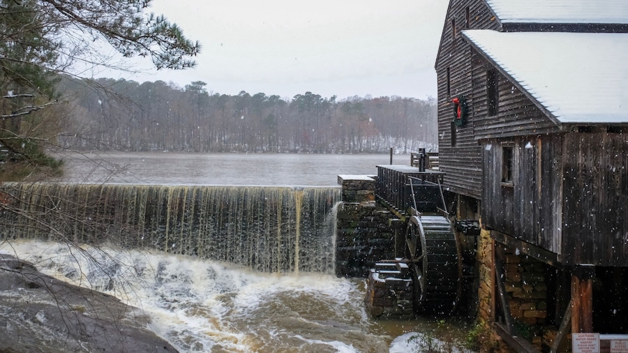 A festive view of the old rustic water mill and waterfall as snow falls at Historic Yates Mill County Park during the Christmas season in Raleigh North Carolina.