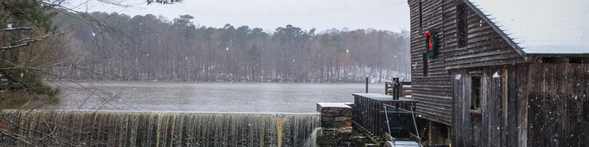 A festive view of the old rustic water mill and waterfall as snow falls at Historic Yates Mill County Park during the Christmas season in Raleigh North Carolina.
