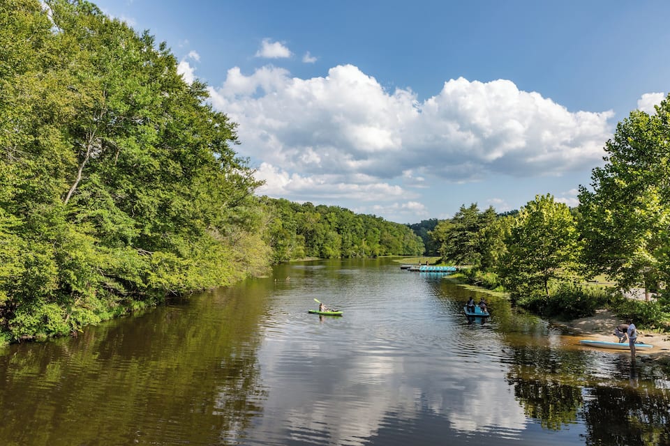 A river surrounded by dense green trees, a person paddling a green kayak, and a small boat with people on the shore.