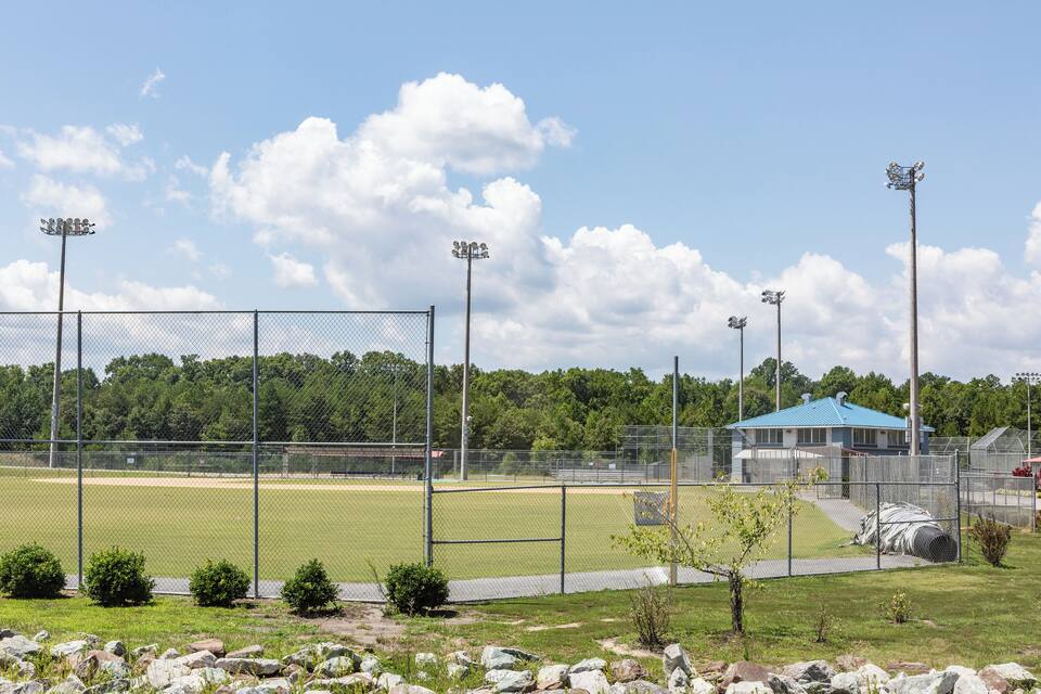 Um campo de beisebol com cercado de arame, holofotes, um edifício com telhado azul e uma área arborizada ao fundo.