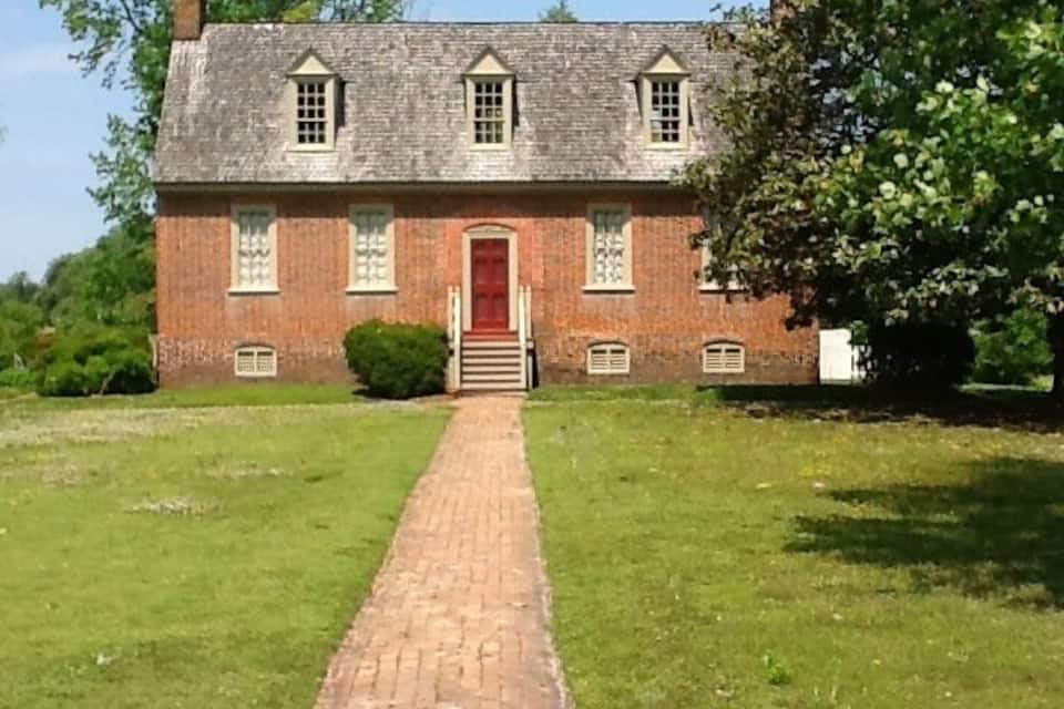 Un bâtiment en briques avec une porte rouge, un chemin en briques menant à l’entrée et un arbre sur la droite.