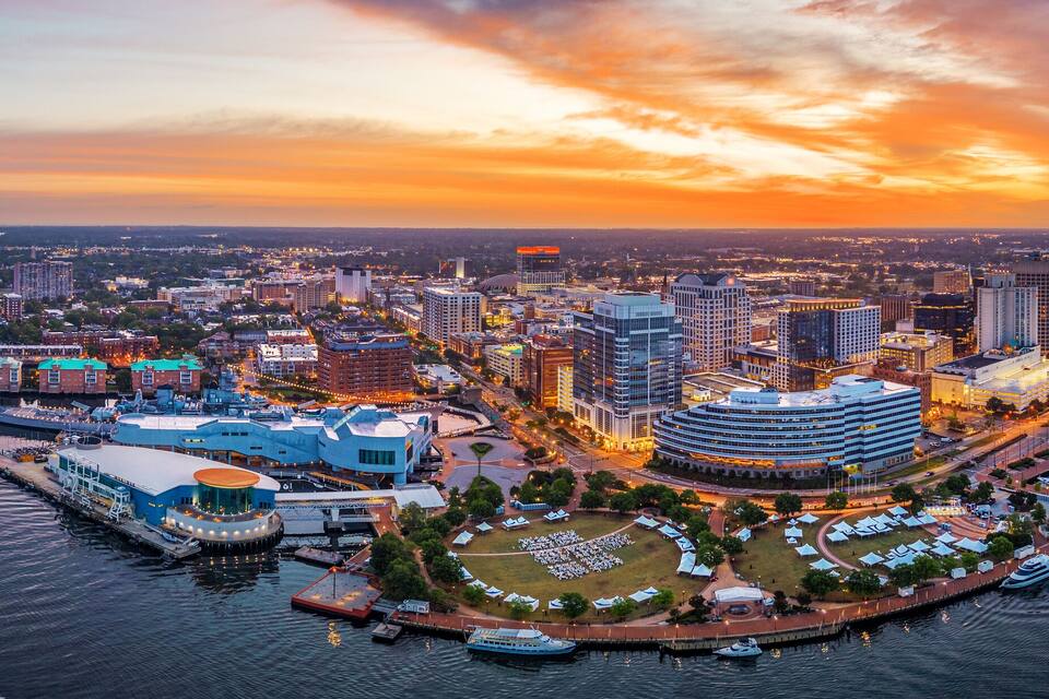 A waterfront cityscape at sunset with a river, buildings, and a bridge.