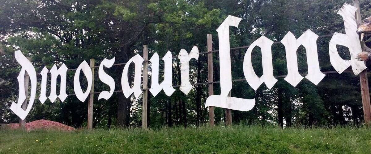 A sign reading "Dinosaur Land" with a backdrop of trees and grass.
