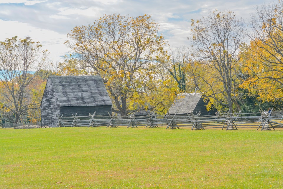 A rustic barn surrounded by a wooden fence, with autumn-colored trees in the background.