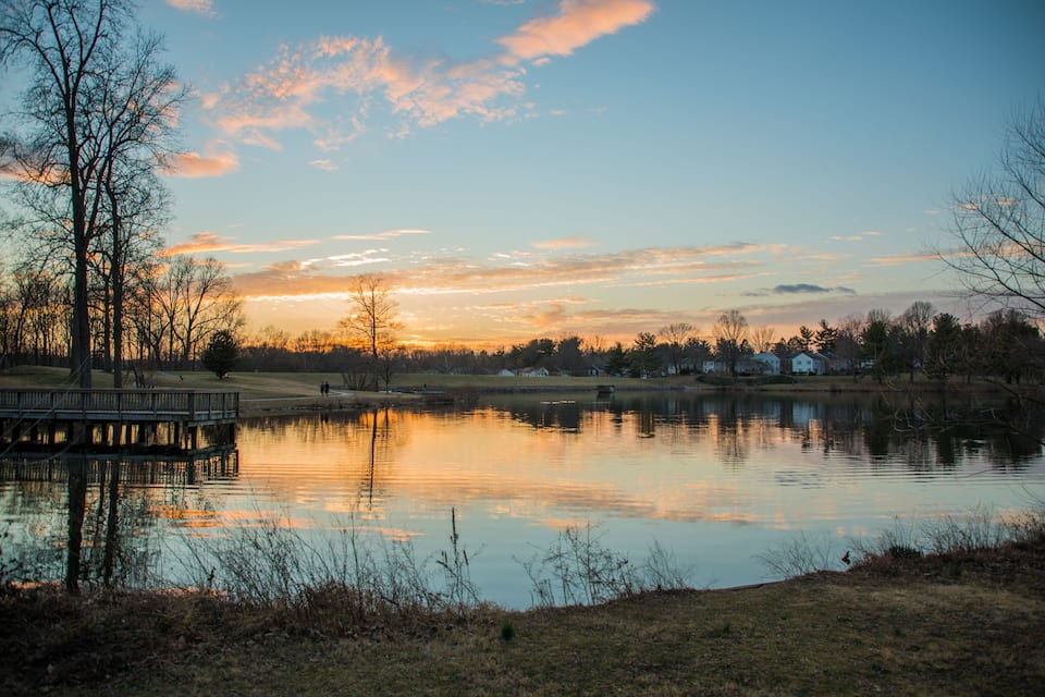 Un lago tranquilo al atardecer con un muelle de madera, rodeado de árboles y casas.