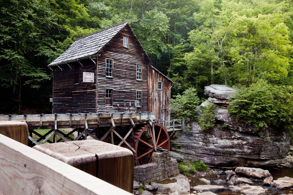 A wooden mill house with a water wheel on a rocky riverbank surrounded by dense forest.