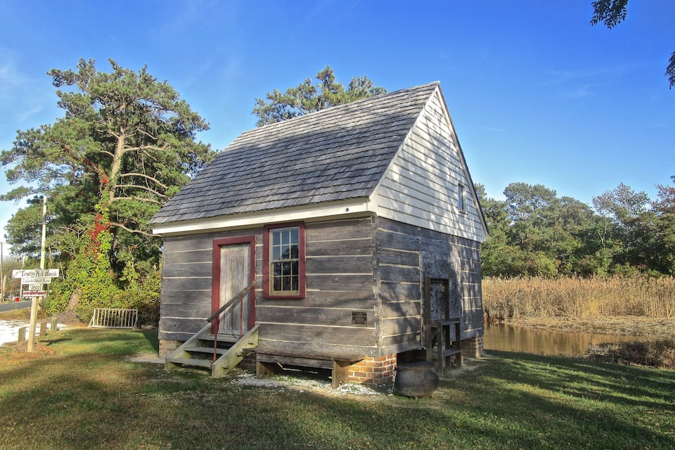 A small wooden house with a red door and a chimney, surrounded by grass and trees.