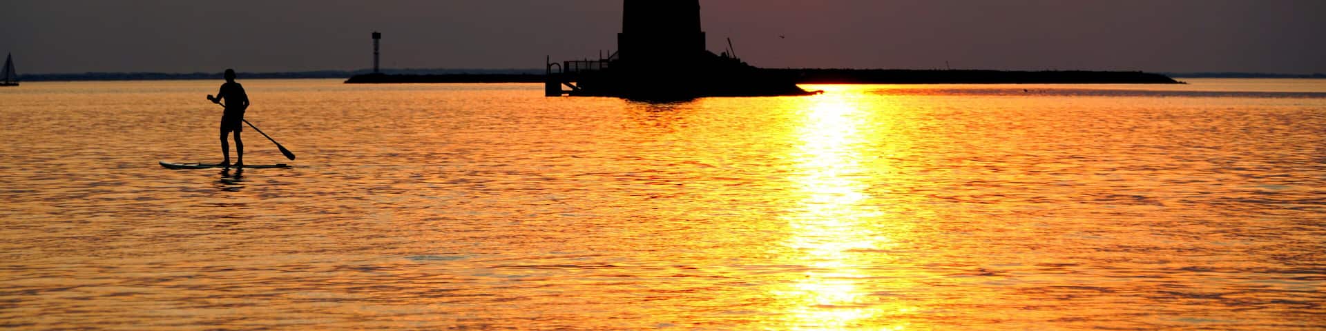 Silhouette of a light house and a man on a paddle board during sunset at Cape Henlopen State Park, Lewes, Delaware, U.S.A