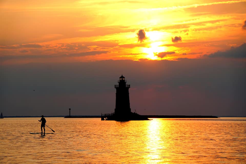 A lighthouse silhouette against a sunset, a person paddleboarding, and calm waters.