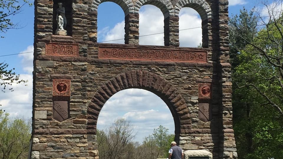 A stone archway with inscriptions and statues, surrounded by grass and trees.