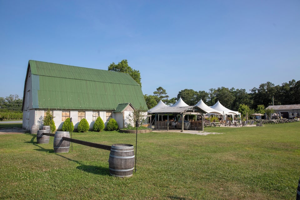 A barn with a green roof, white walls, and several barrels in a grassy field.