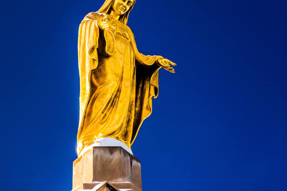 A golden statue of a robed figure with outstretched arms, set against a clear blue sky.