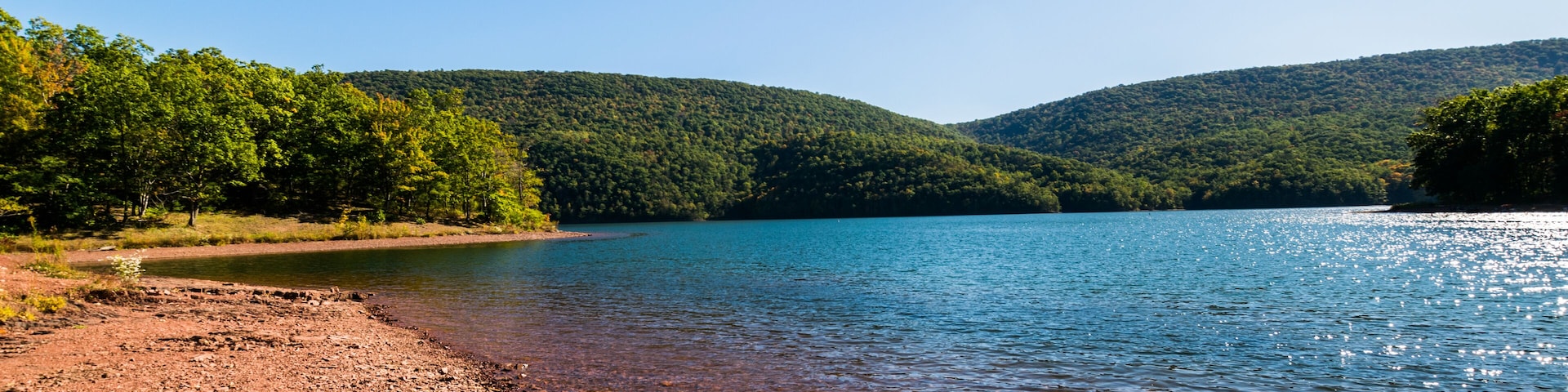 A clear lake with a rocky shoreline and forested hills in the background.