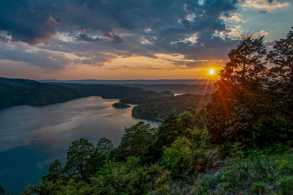 A sunset over a calm lake surrounded by forested hills.