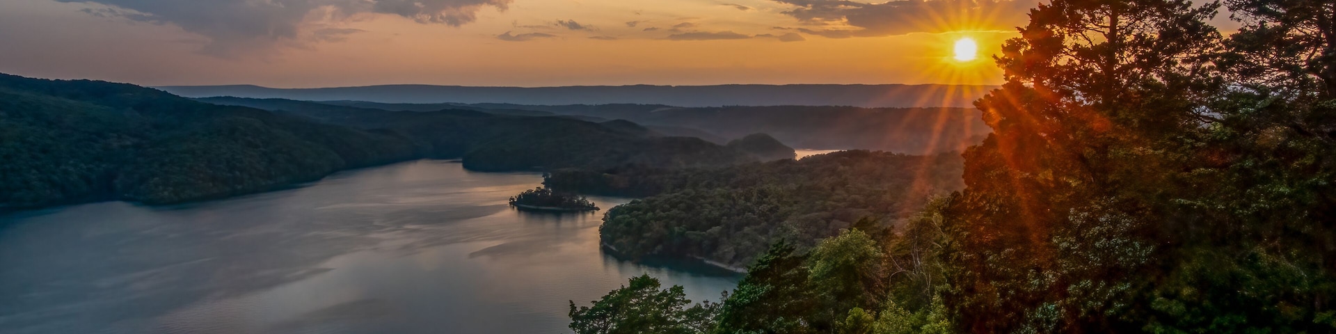 Ein Sonnenuntergang über einem ruhigen See, umgeben von bewaldeten Hügeln.