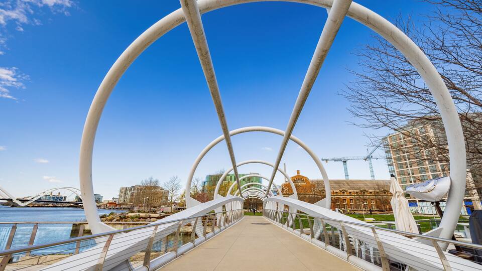 A pedestrian bridge with white arches over a river, with buildings and a crane in the background.