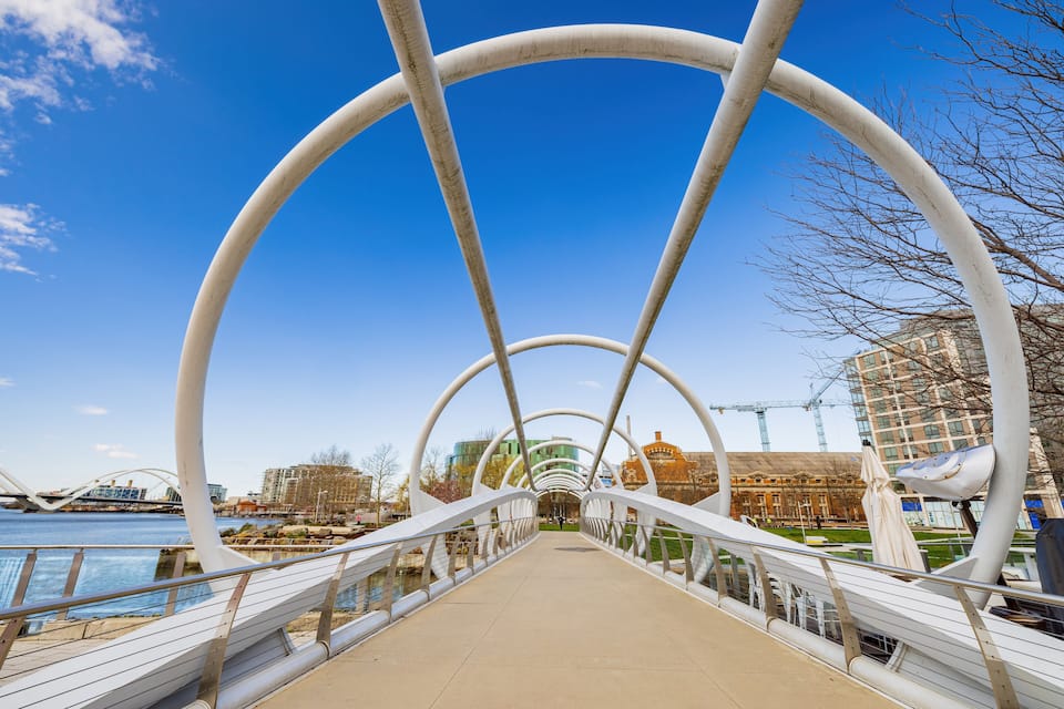 A pedestrian bridge with white arches over a river, with buildings and a crane in the background.