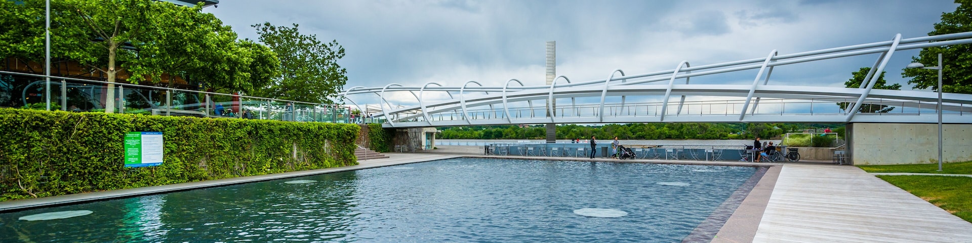 A swimming pool with a wooden deck, surrounded by greenery and a modern bridge structure.
