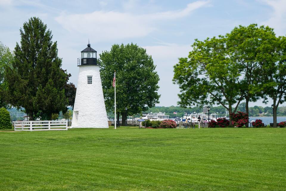 Un phare blanc coiffé d’une partie noire, entouré d’une végétation luxuriante, avec de l’herbe verte et des arbres.