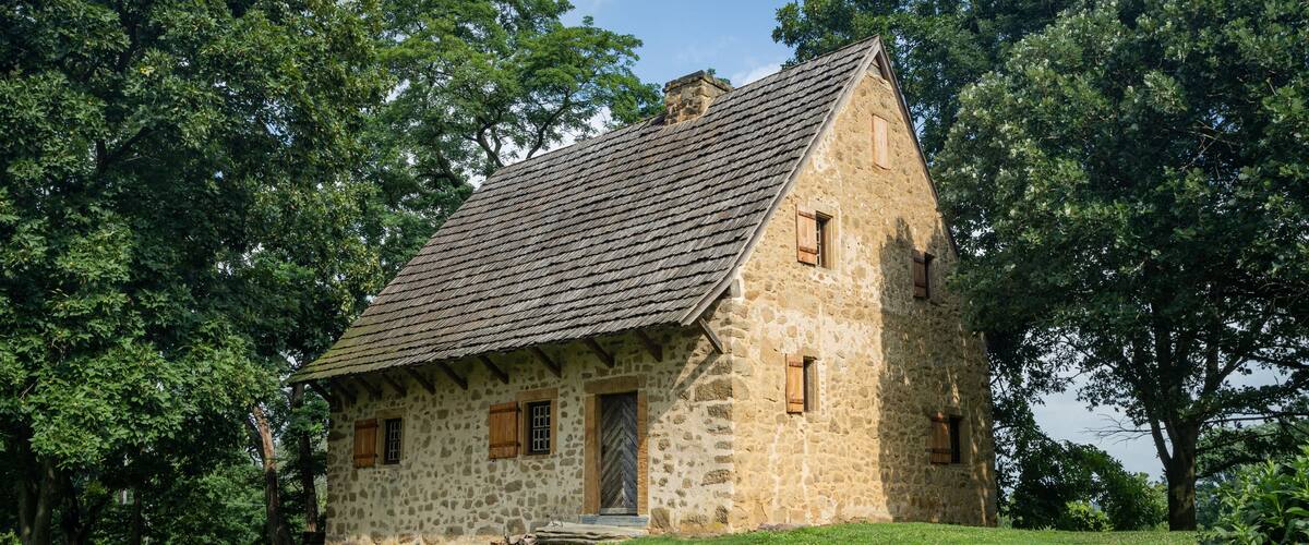A stone house with a steep gabled roof, surrounded by lush green trees.