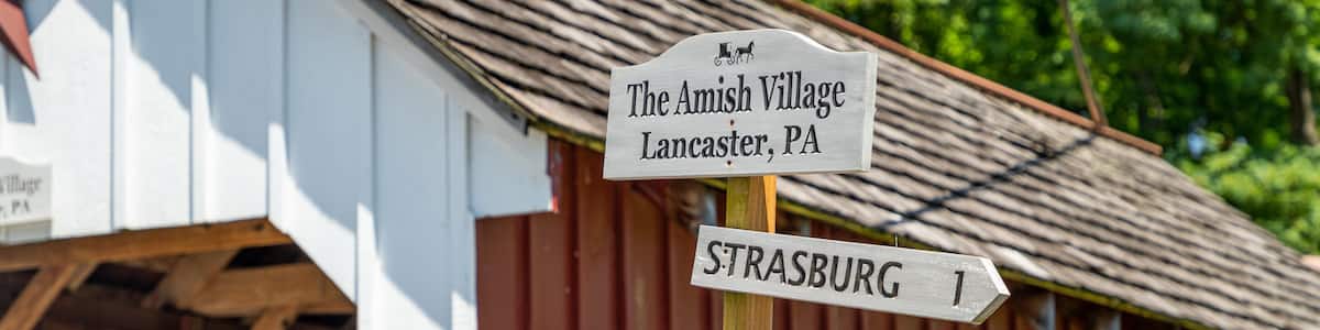 A wooden signpost with directions to The Amish Village, Strasburg, Amish Wares, and Paradise.